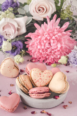 Heart shaped sweet macarons and flowers in pastel colors on a pink background. A delicious dessert or gift for Valentine's Day or Mother's Day. Vertical.