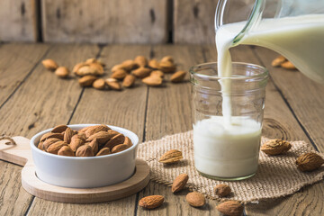 Vegan almond milk pouring into a glass, almond kernels and whole almonds on the old rustic wooden...