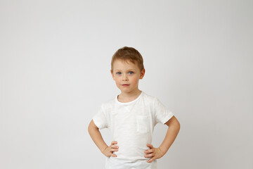 A boy with blue eyes of Caucasian nationality looks confidently into the camera. The child is dressed in a white T-shirt and stands with his hands on his hips.