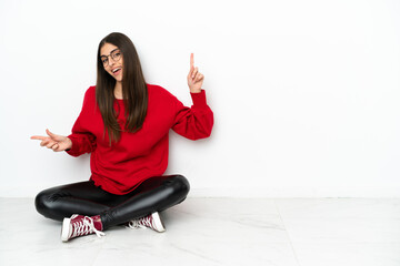 Young woman sitting on the floor isolated on white background pointing finger to the laterals and happy