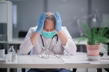 During the pandemic, an elderly male doctor is very tired from work, he sits at his desk in the resident's office and holds his head