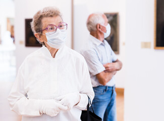 Obraz premium elderly European woman in mask protecting against covid examines paintings on display in hall of art museum