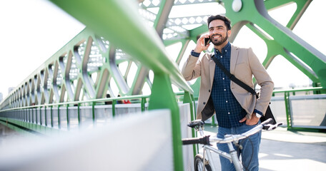 Young business man commuter with bicycle going to work outdoors in city, using smartphone.