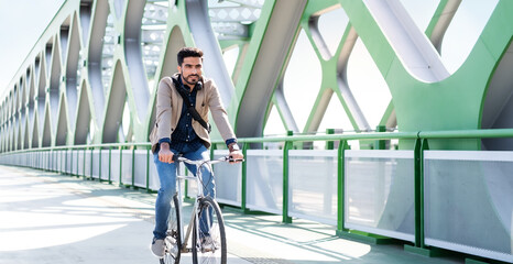 Young business man commuter with bicycle going to work outdoors in city, riding on bridge.