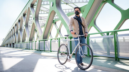 Young business man commuter with bicycle going to work outdoors on bridge in city, coronavirus...