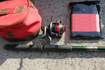 Close-up of a fishing rod with a red reel and colorful line, a fishing bag and fish bait in a rectangular plum-flavored package