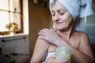 Obraz na płótnie Canvas Happy senior woman wrapped in a towel in bathroom at home, applying homemade body moisturiser.