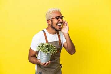 Young Colombian man holding a plant isolated on yellow background shouting with mouth wide open to...