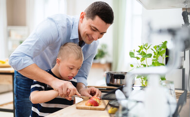 Father with happy down syndrome son indoors in kitchen, chopping apple.