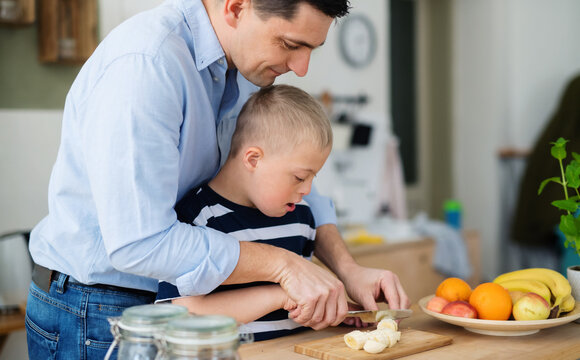 Father with happy down syndrome son indoors in kitchen, chopping fruit.