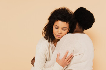 african american adult daughter with closed eyes embracing with middle aged mother isolated on beige
