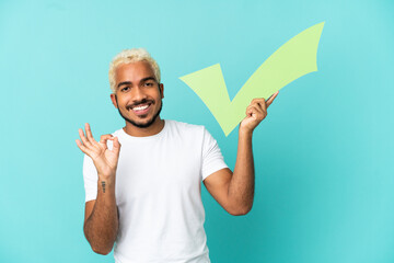 Young Colombian handsome man isolated on blue background holding a check icon and doing OK sign