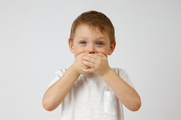 A small boy with blue and cheerful eyes on a white background covered his mouth with his hands. The child holds back the emotions of joy by covering his mouth with his hands