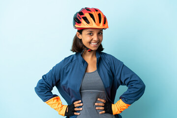 Young cyclist woman isolated on blue background posing with arms at hip and smiling - obrazy, fototapety, plakaty