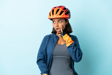 Young cyclist woman isolated on blue background whispering something with surprise gesture while looking to the side - obrazy, fototapety, plakaty