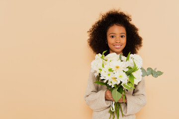 Obraz na płótnie Canvas smiling african american preteen girl with bouquet of flowers in hands isolated on beige