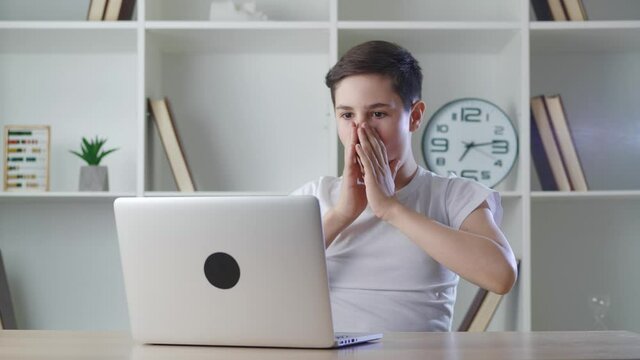 Shocked 13-year-old boy rejoices at the achievements of working at a computer at home. Teenager is pleasantly surprised by the sudden victory while playing with his laptop.