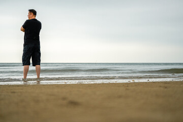 A tall Asian man stands on the beach, close-up back view