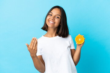 Young asian girl holding an orange isolated on blue background inviting to come with hand. Happy that you came