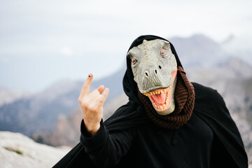 Caucasian man with a dinosaur mask and a cape making the sign of the horns with his hand in the...
