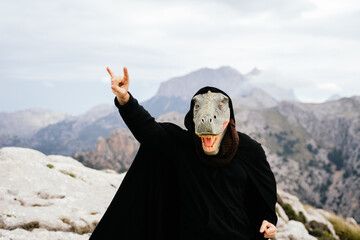 Caucasian man with a dinosaur mask and a cape making the sign of the horns with his hand in the...