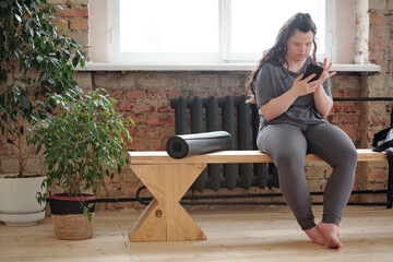Restful female with Down syndrome scrolling in smartphone while sitting on bench after workout