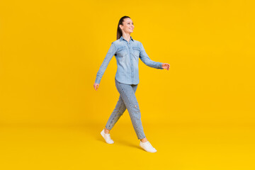 Fototapeta na wymiar Full length photo of excited sweet young lady dressed jeans shirt walking looking empty space smiling isolated yellow color background