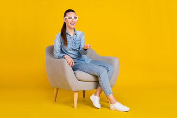 Photo of pretty shiny young lady dressed jeans shirt spectacles sitting chair looking empty space smiling isolated yellow color background