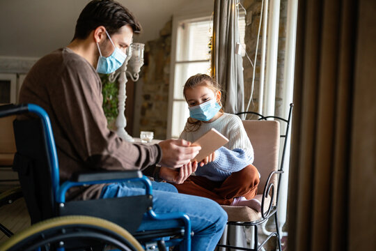 Father with disability in wheelchair using tablet at home with child while wearing masks.
