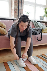 Young female with Down syndrome in sportswear getting ready for yoga training while sitting on sofa at home