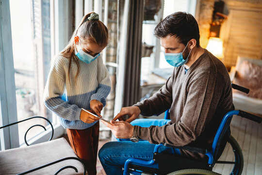 Father with disability in wheelchair using tablet at home with child while wearing masks.