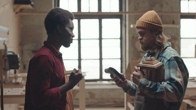 Medium shot of young African man paying bearded Caucasian joiner using contactless credit card with NFC technology, then taking handmade wooden lantern and shaking hand of carpenter