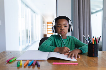 African american boy in online school class, using headphones and writing in his notebook