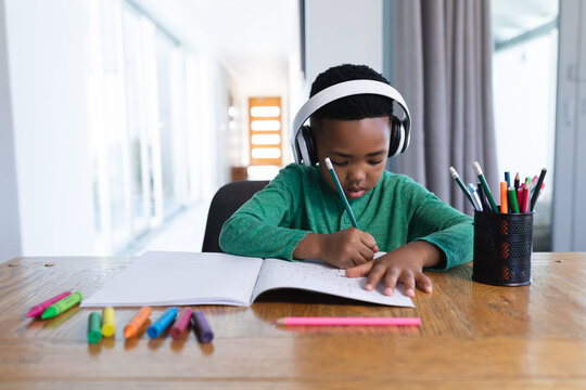 African american boy in online school class, using headphones and writing in his notebook