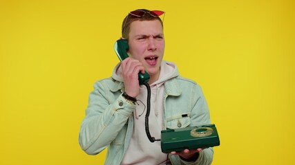 Crazy sincere teen student boy 20s years old in denim jacket talking on wired vintage telephone of 80s, fooling, making silly faces. Young funny adult man isolated on yellow background