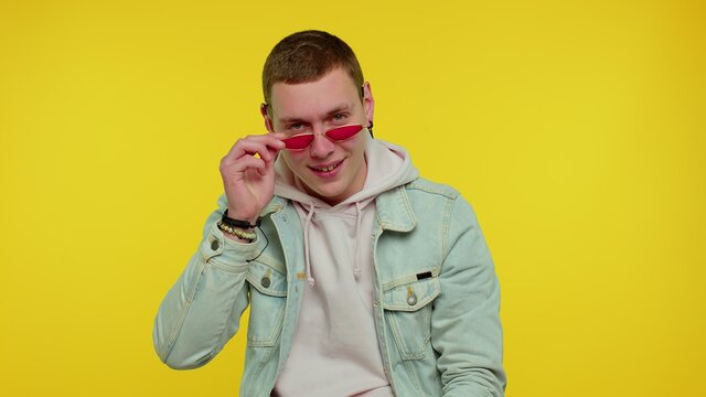 Playful Happy Teenager Student Boy 18 Years Old Wearing Sunglasses Blinking Eye Looking At Camera With Toothy Smile, Winking And Flirting Expressing Optimism. Young Adult Man On Yellow Wall Background