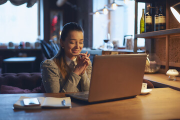 Fototapeta premium Young beautiful woman sits at a wooden table in a cafe indoors and works on a laptop. Conception of remote work and freelance work.