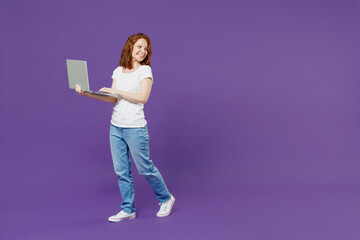 Full length young fun happy satisfied redhead woman wear white basic casual t-shirt holding using laptop pc computer chat online look aside isolated on dark violet color background studio portrait.