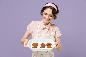 Young smiling housewife housekeeper chef cook baker woman wearing pink apron show chocolate cookies biscuits on white baking form isolated on pastel violet background. Cooking food process concept