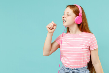 Little redhead kid girl 12-13 years old wearing pink striped t-shirt headphones listen to music sing song in microphone isolated on pastel blue background studio. Children lifestyle childhood concept.