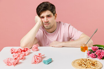 Sick ill allergic man has red watery eyes runny stuffy sore nose suffer from allergy trigger symptom hay fever sits with napkin plant allergen products isolated on pastel pink color background studio