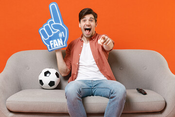 Young man football fan in shirt foam glove finger support favorite team with soccer ball sit home...