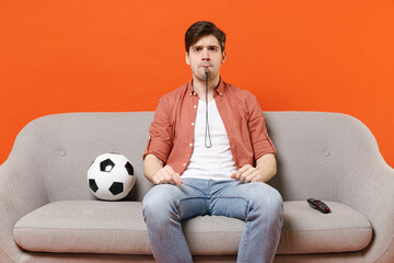 Young serious man football fan wear shirt support team with soccer ball sit on sofa at home watching tv live stream blow whistle isolated on orange background People sport leisure lifestyle concept