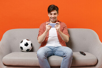 Young gambling man football fan wear shirt support team with soccer ball sit on sofa home watch tv use play racing on mobile cell phone smartphone isolated on orange background. People sport concept © ViDi Studio