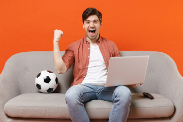 Young fun excited man football fan in shirt support team with soccer ball sit on sofa home watch tv...