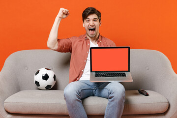 Young fun man football fan in shirt support team with soccer ball sit sofa home watch tv use laptop...