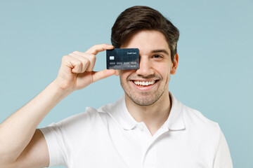 Young happy smiling cheerful fun caucasian man 20s wearing white casual basic t-shirt covering eye with credit bank card isolated on pastel blue background studio portrait People lifestyle concept