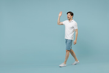 Fototapeta na wymiar Full length young fun happy unshaven cheerful caucasian man 20s wear white basic t-shirt walking go waving hand say hello isolated on pastel blue background studio portrait. People lifestyle concept.