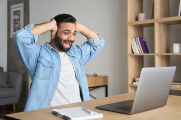 Obraz premium Overjoyed happy indian latin guy, lucky winner, sitting at desk watching virtual lottery results on laptop holding his head with hands. Fortunate young trader specialist got stock market job.