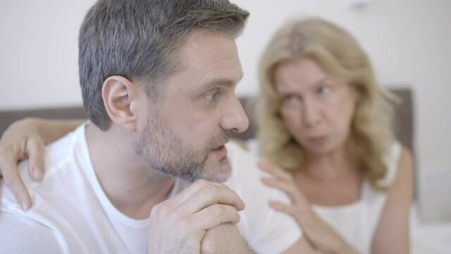Middle-aged woman trying to talk to offended husband sitting in bed, problems